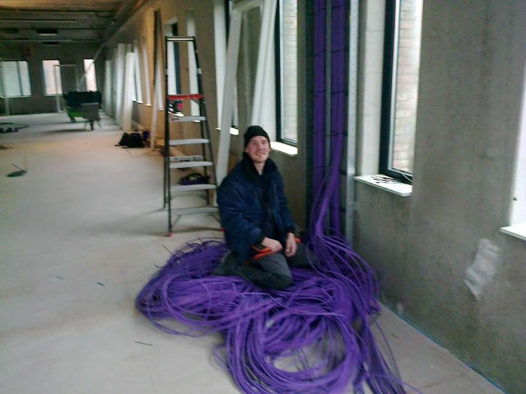 Uśmiechnięty elektryk siedzi na stercie fioletowych kabli w surowym wnętrzu budynku w trakcie instalacji elektrycznej, drabina oparta o ścianę w tle.