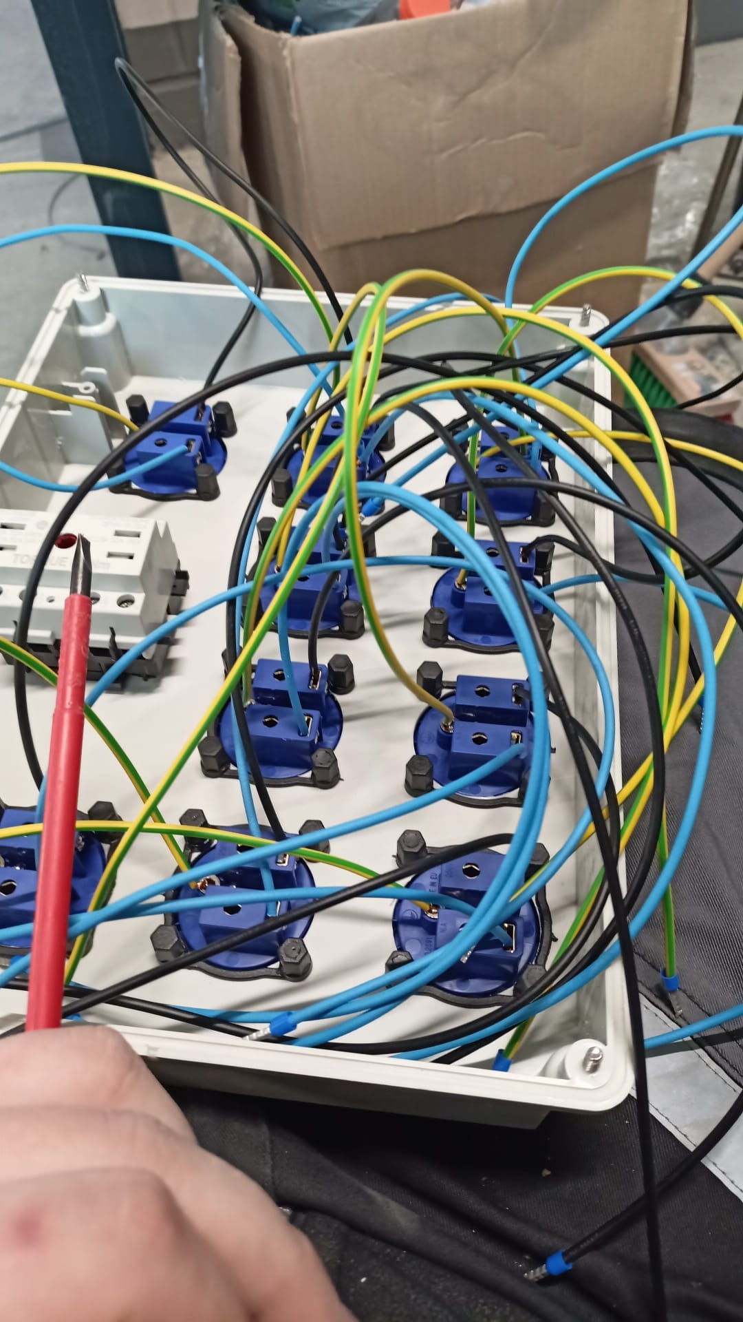 Rozdzielnica elektryczna w trakcie montażu: widoczne niebieskie gniazda, przewody w izolacji żółto-zielonej, niebieskiej i czarnej, oraz czerwony próbnik napięcia w dłoni montera.