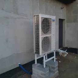 Zewnętrzna jednostka pompy ciepła Panasonic, zamontowana na betonowej podstawie przy surowej ścianie budynku, z widocznym niebieskim wężem odprowadzającym skropliny.