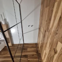 Nowoczesna łazienka z kabiną prysznicową typu walk-in z czarnymi profilami, drewnopodobne płytki na podłodze i ścianie, białe płytki na pozostałych ścianach, widoczny odpływ liniowy.