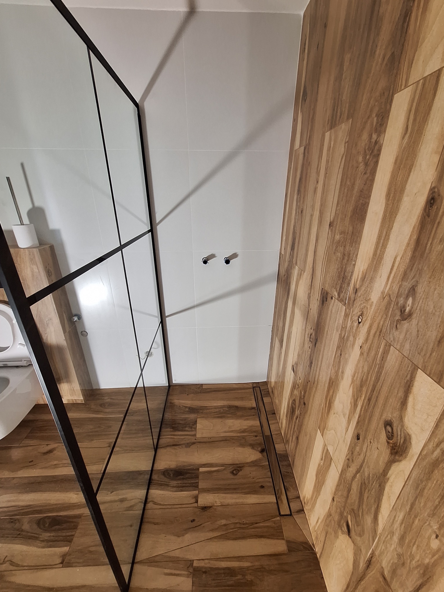 Nowoczesna łazienka z kabiną prysznicową typu walk-in z czarnymi profilami, drewnopodobne płytki na podłodze i ścianie, białe płytki na pozostałych ścianach, widoczny odpływ liniowy.