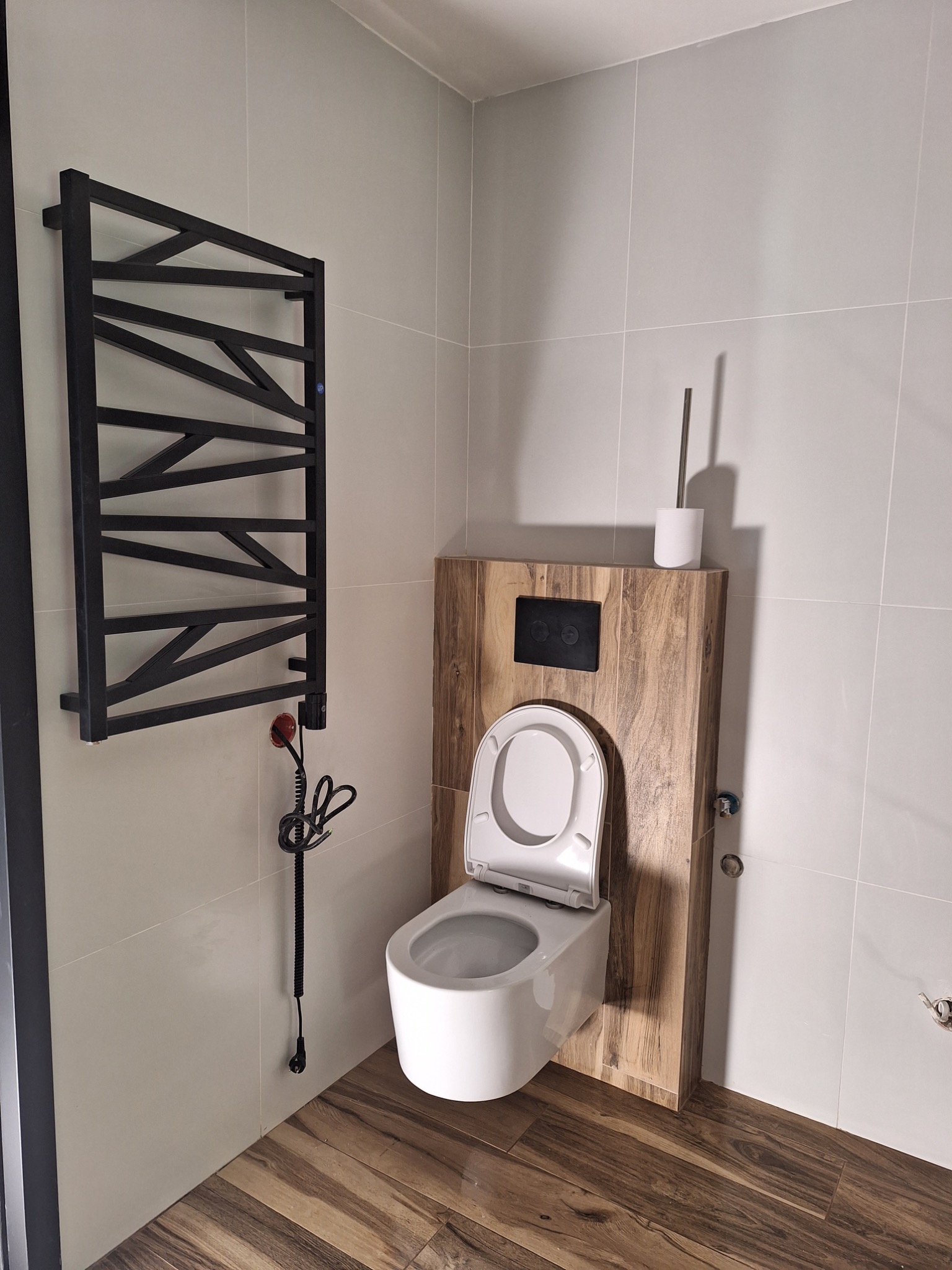 Nowoczesna łazienka z podwieszaną toaletą na drewnianym stelażu, czarnym grzejnikiem drabinkowym i płytkami imitującymi drewno na podłodze.