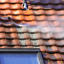 Czyszczenie czerwonej dachówki z mchu i zabrudzeń za pomocą myjki ciśnieniowej, widoczny fragment okna dachowego.