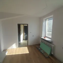 Pokój w trakcie prac przygotowawczych: świeżo pomalowane ściany na biało, drewniana podłoga, grzejnik pod oknem, płyty gipsowo-kartonowe oparte o ścianę, widoczne kable wystające z sufitu.