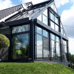 Luk’s & Roses Glas u. Wintergärtenfabrik - Nowoczesna zabudowa tarasu z czarnymi ramami i dużymi przeszkleniami, widoczna drabina oparta o ścianę, trawnik i osoba siedząca na trawie.