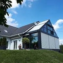 Luk’s & Roses Glas u. Wintergärtenfabrik - Zadaszony taras w Wien z nowoczesną konstrukcją i roletami przeciwsłonecznymi, widok z zewnątrz na tle zieleni i błękitnego nieba.