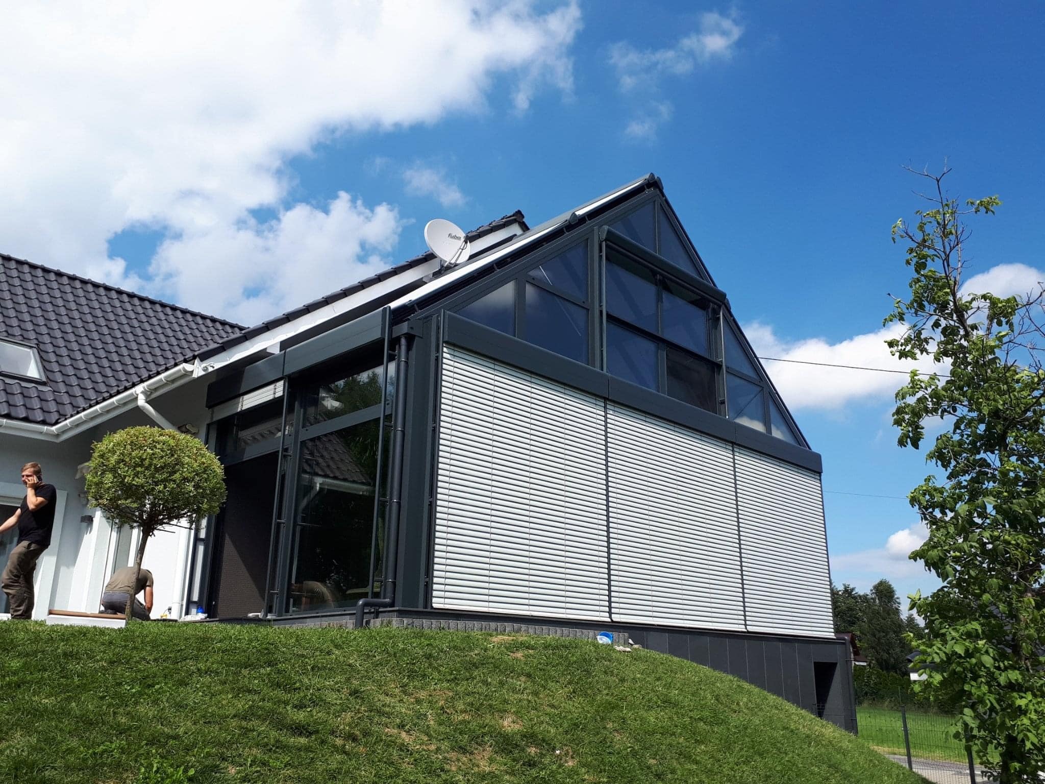 Nowoczesna zabudowa balkonu z przeszkleniami i żaluzjami zewnętrznymi, widok z zewnątrz na tle zielonego trawnika i błękitnego nieba z chmurami.