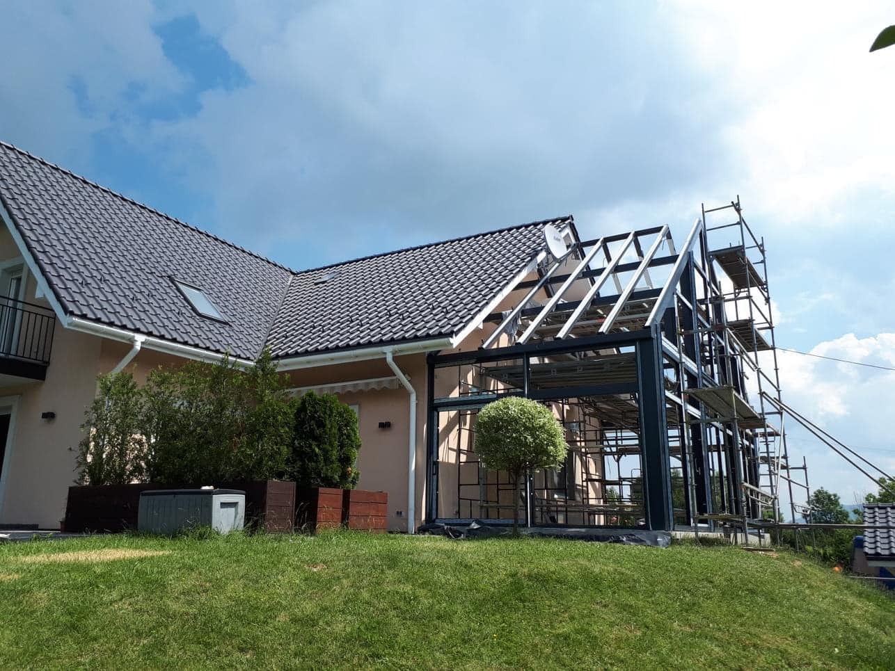 Konstrukcja szklarni przylegającej do domu, widoczne rusztowania, elementy stalowe i szklane, trawnik z przodu.