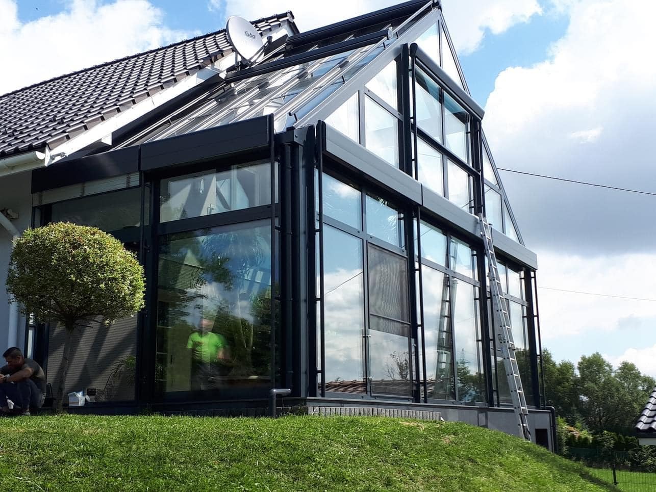 Nowoczesna zabudowa tarasu z czarnymi ramami i dużymi przeszkleniami, widoczna drabina oparta o ścianę, trawnik i osoba siedząca na trawie.