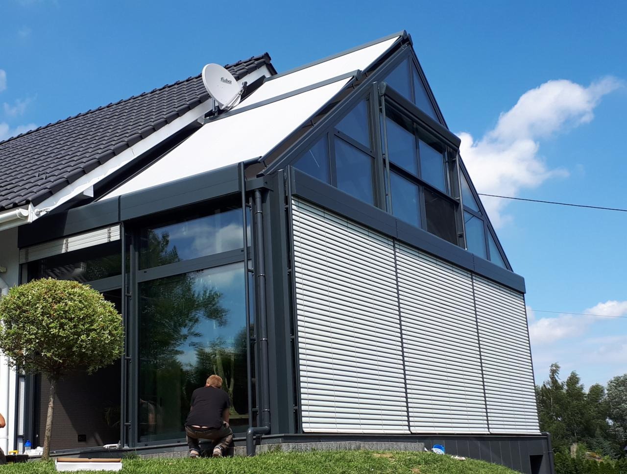 Nowoczesna zabudowa tarasu z aluminiową konstrukcją, żaluzjami zewnętrznymi i skośnym dachem, widoczna osoba w trakcie montażu.