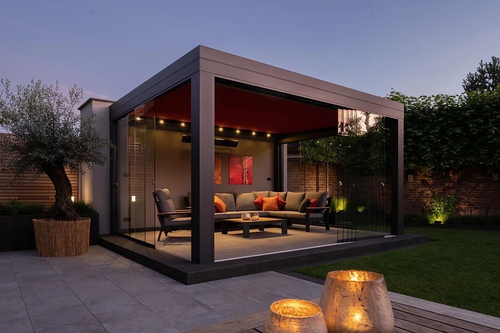 Nowoczesna szara pergola z przesuwnymi szklanymi ścianami, oświetleniem LED i czerwonym zadaszeniem, na kamiennym tarasie z drzewkiem oliwnym w plecionym koszu i świecami w lampionach.