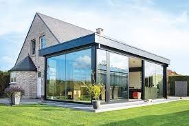 Szklana zabudowa tarasu przylegająca do domu z cegły, widok z zewnątrz, zieleń trawnika w pierwszym planie.