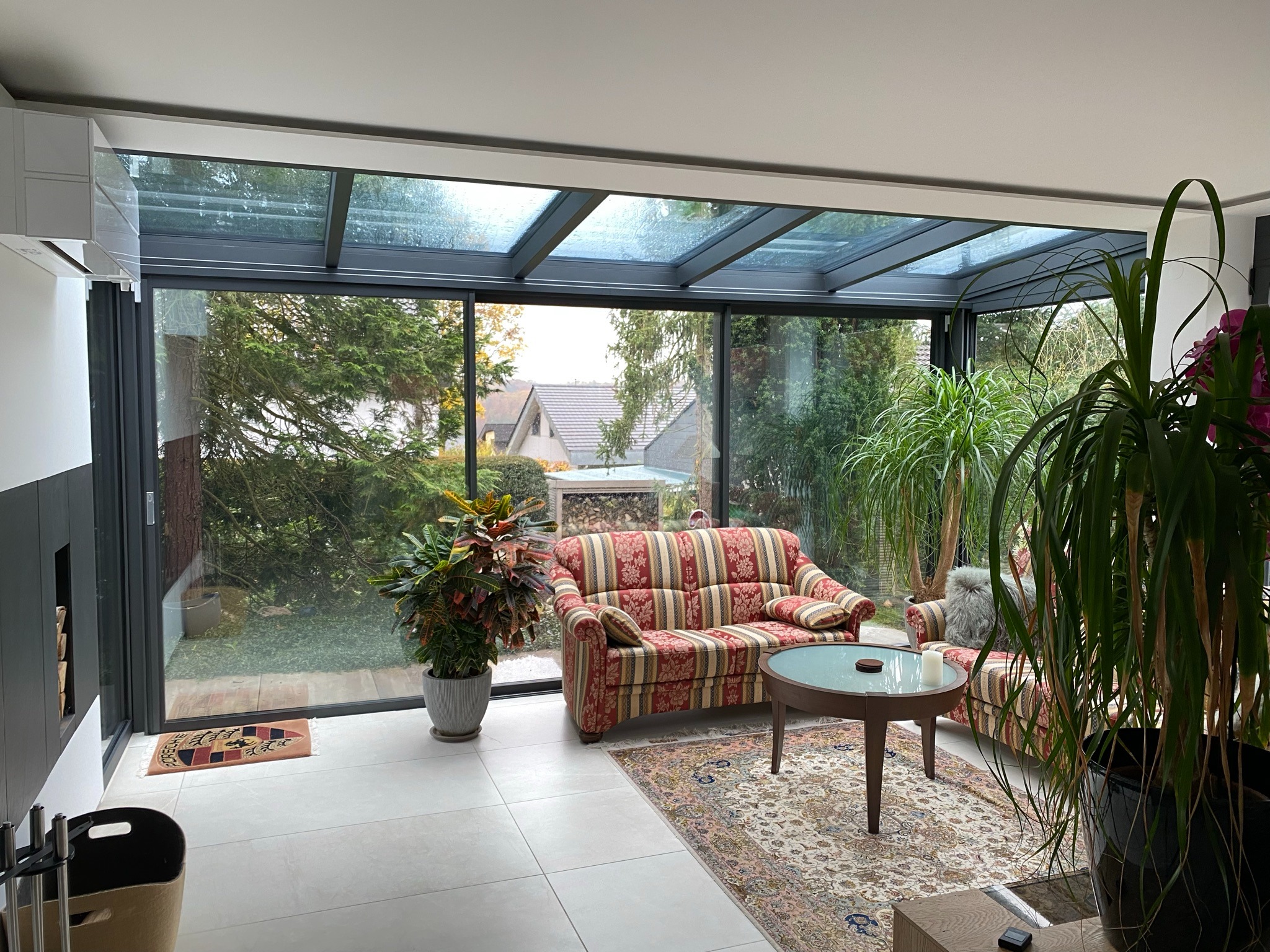 Nowoczesna zabudowa balkonu z przeszklonym dachem i ścianami, widoczne meble wypoczynkowe i rośliny doniczkowe, na zewnątrz widok na ogród.