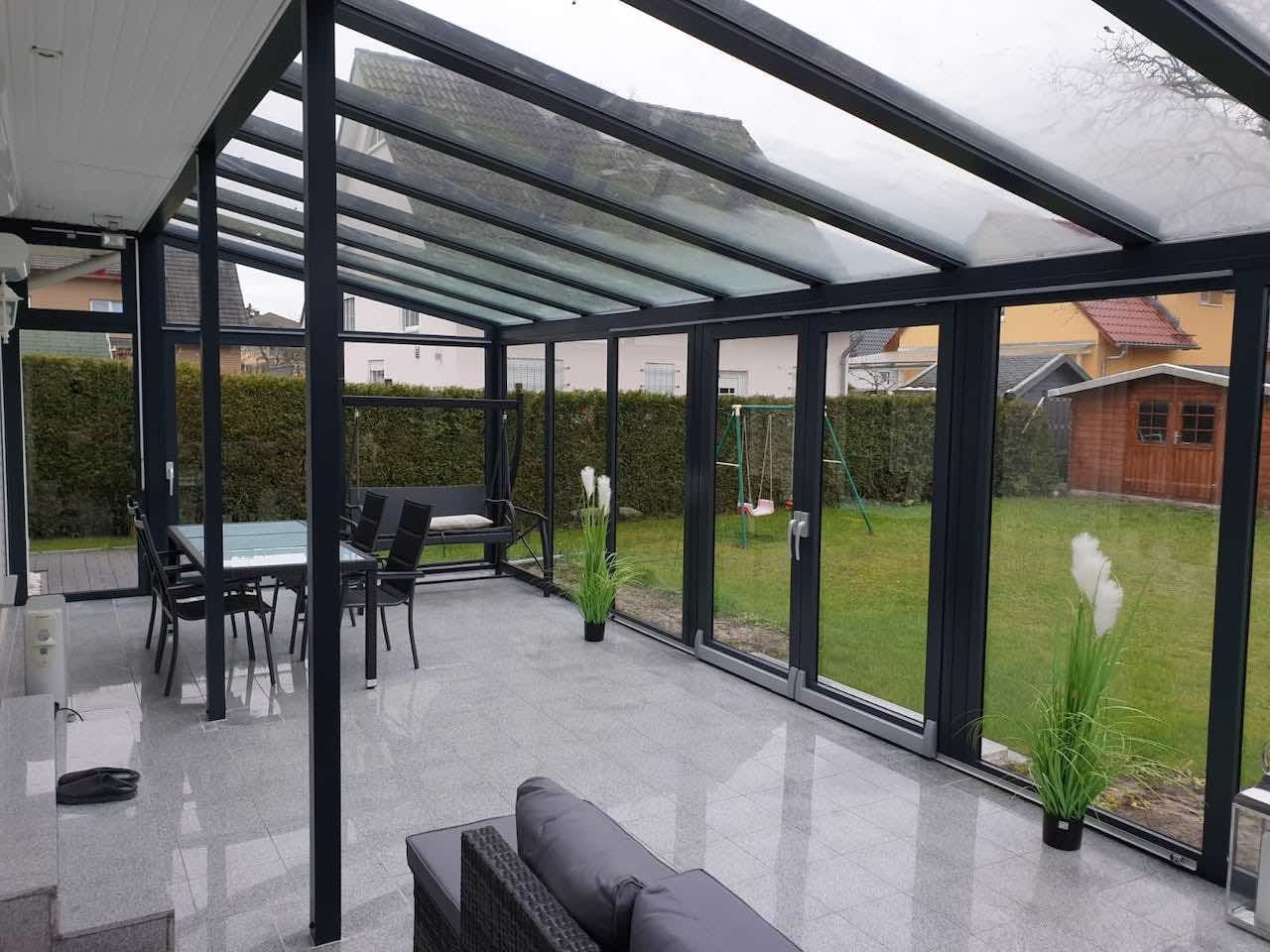 Szklana zabudowa tarasu z czarnymi profilami, widok na ogród z huśtawką i domkiem narzędziowym, podłoga wyłożona jasnoszarymi płytkami, wewnątrz stół z krzesłami i kanapa.