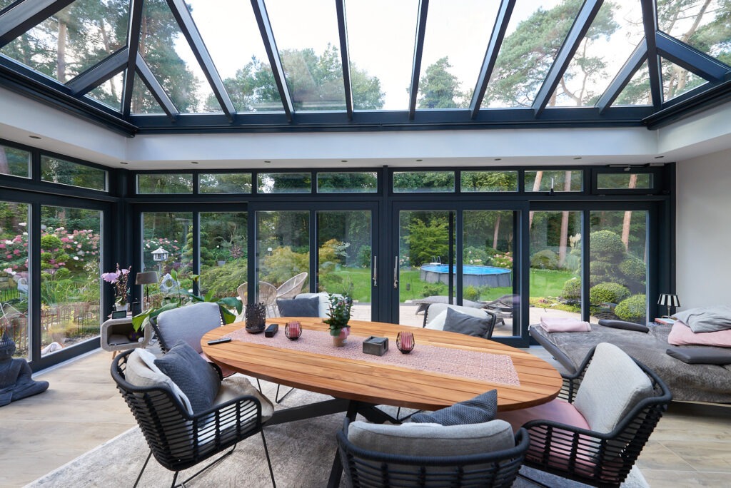 Przestronna oranżeria z aluminiową konstrukcją, przeszklonym dachem i przesuwnymi drzwiami, widok na zadbany ogród z basenem.