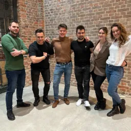 Grupa sześciu młodych osób, uśmiechniętych i stojących blisko siebie na tle ściany z czerwonej cegły i przeszklonej ściany z metalowymi ramami, w tle widoczne półki z butelkami.