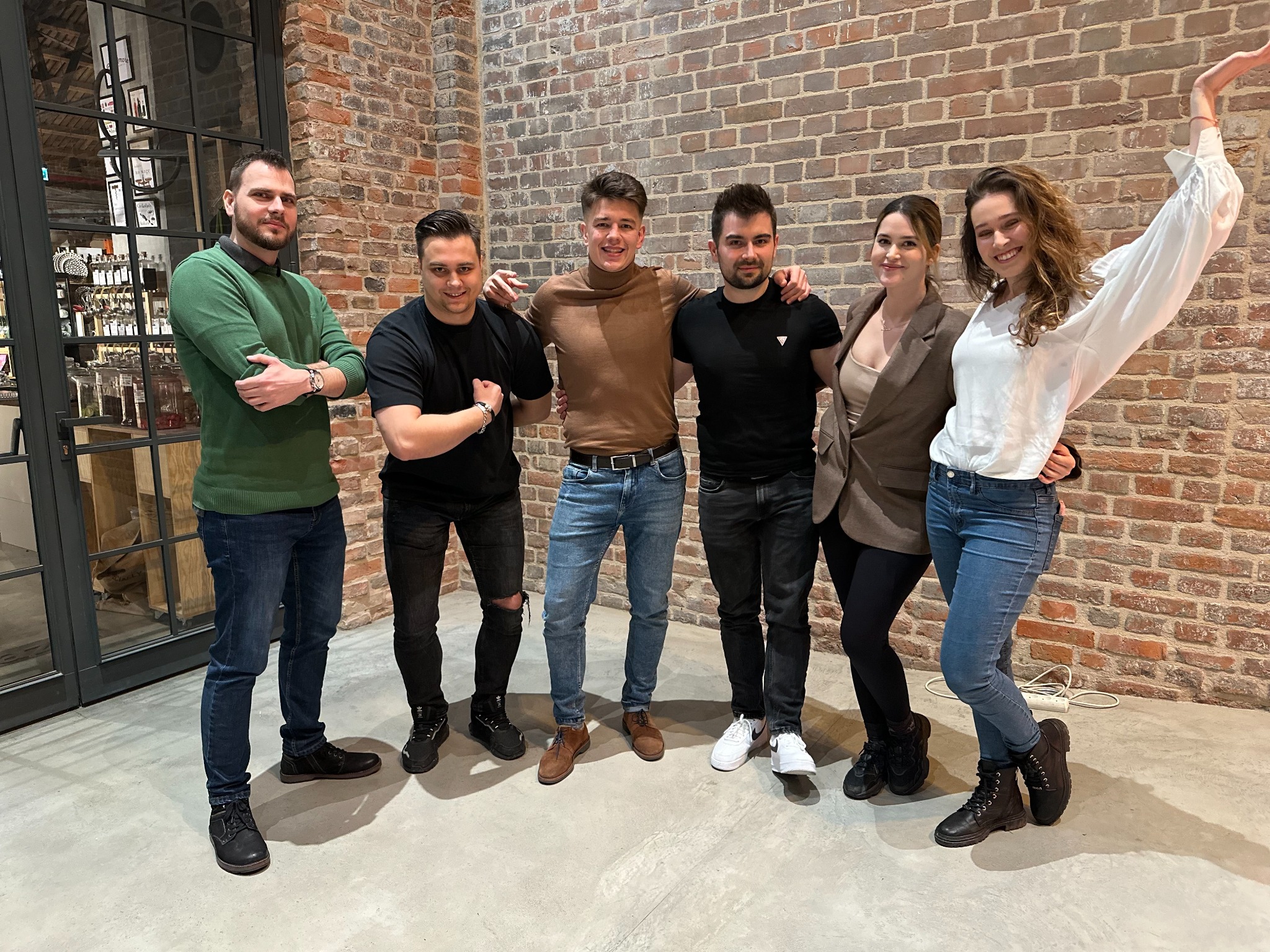 Grupa sześciu młodych osób, uśmiechniętych i stojących blisko siebie na tle ściany z czerwonej cegły i przeszklonej ściany z metalowymi ramami, w tle widoczne półki z butelkami.
