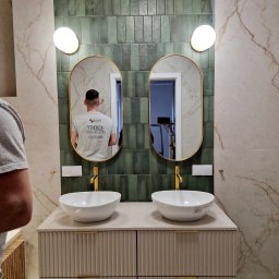 DP REM-BUD DANIEL PŁACHECKI - Wykończona łazienka z dwoma umywalkami nablatowymi, złotymi bateriami i owalnymi lustrami podświetlonymi lampami. Na ścianach płytki imitujące marmur i zielone pionowe płytki. W lustrze odbija się...