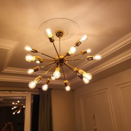 DP REM-BUD DANIEL PŁACHECKI - Złoty, nowoczesny żyrandol z wieloma żarówkami na tle białego sufitu z dekoracyjnymi listwami i rozetą.