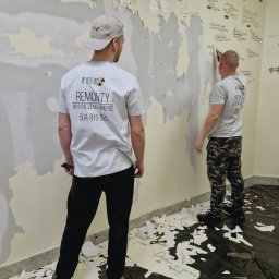 DP REM-BUD DANIEL PŁACHECKI - Dwóch pracowników firmy remontowej w firmowych koszulkach usuwa starą tapetę z pomazanej ściany. Na podłodze folia ochronna i fragmenty oderwanej tapety.