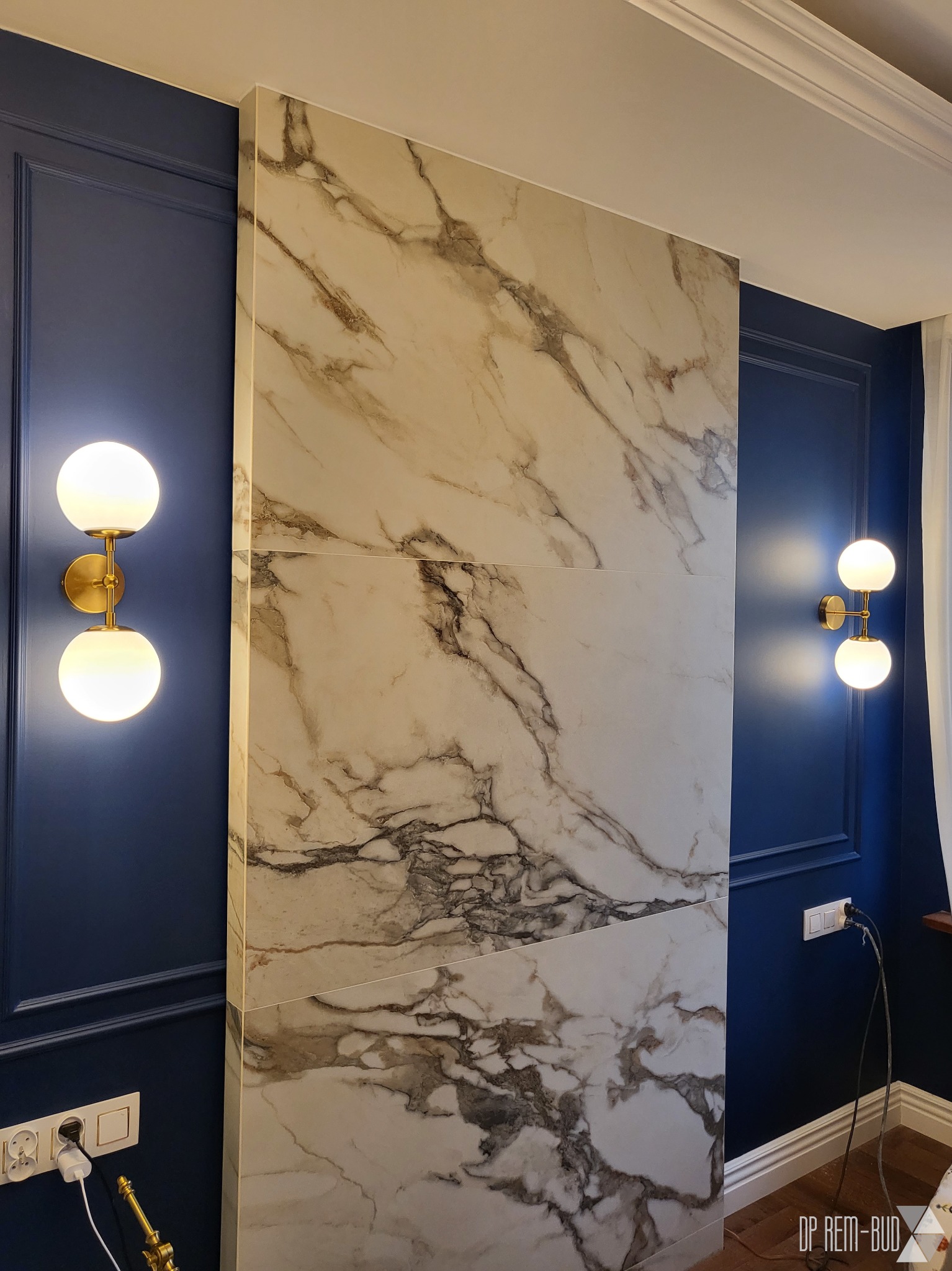 Wykończenie ściany w salonie: płyty imitujące marmur sąsiadują z granatową ścianą z dekoracyjnymi listwami, oświetloną przez dwa nowoczesne kinkiety z kulistymi, mlecznymi kloszami i złotymi...
