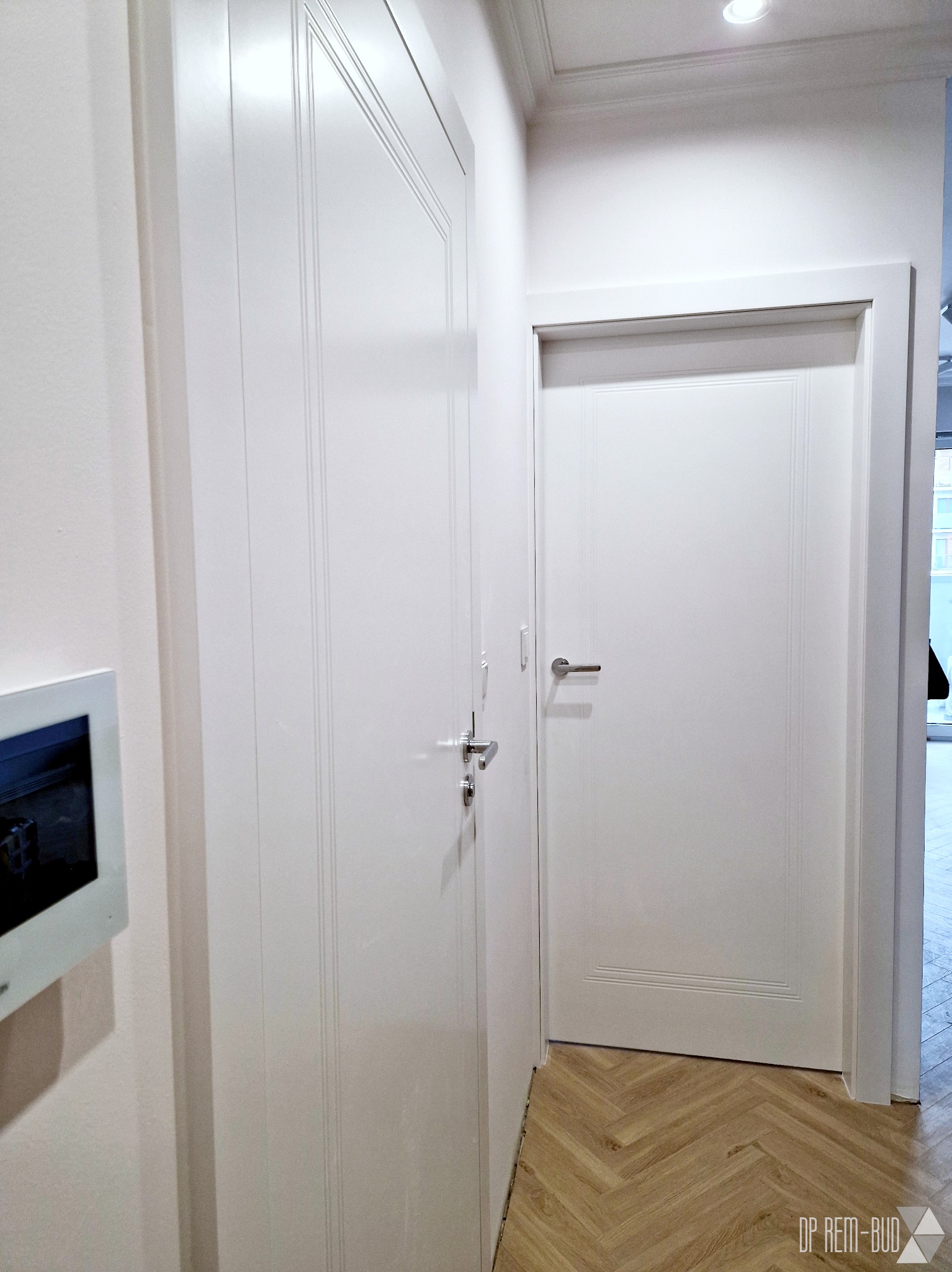 Wykończone wnętrze mieszkania po remoncie: białe, lakierowane drzwi wewnętrzne z frezowanymi zdobieniami, ułożone w jodełkę panele podłogowe oraz panel sterowania inteligentnego domu na ścianie.