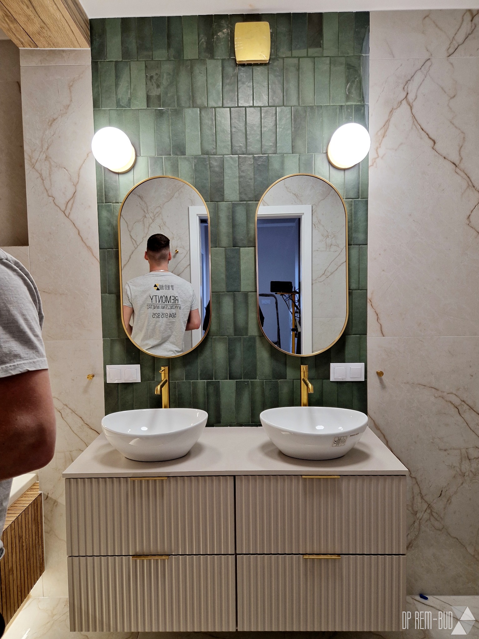 Wykończona łazienka z dwoma umywalkami nablatowymi, złotymi bateriami i owalnymi lustrami podświetlonymi lampami. Na ścianach płytki imitujące marmur i zielone pionowe płytki. W lustrze odbija się...