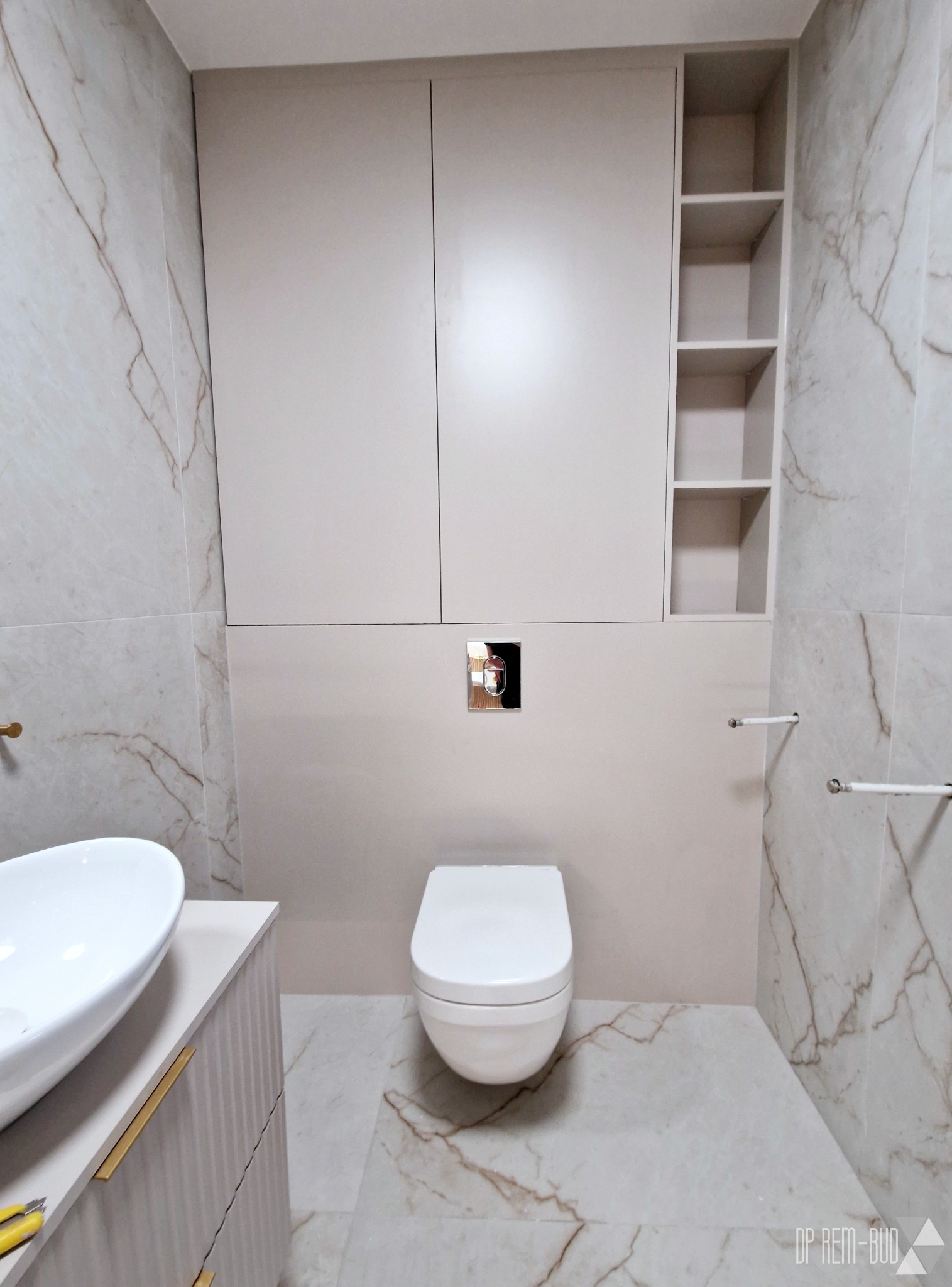 Wykończona łazienka w neutralnych barwach z marmurowymi płytkami, podwieszaną toaletą, szafkami nad nią i umywalką nablatową z szafką w prążki i złotymi uchwytami.