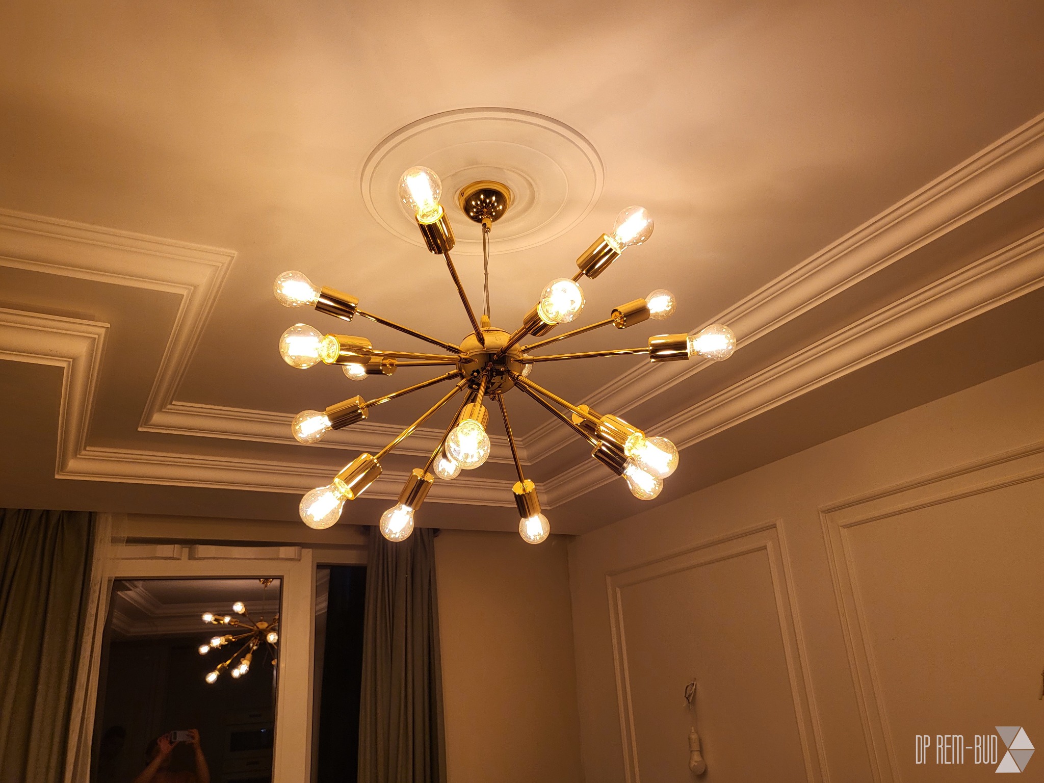 Złoty, nowoczesny żyrandol z wieloma żarówkami na tle białego sufitu z dekoracyjnymi listwami i rozetą.