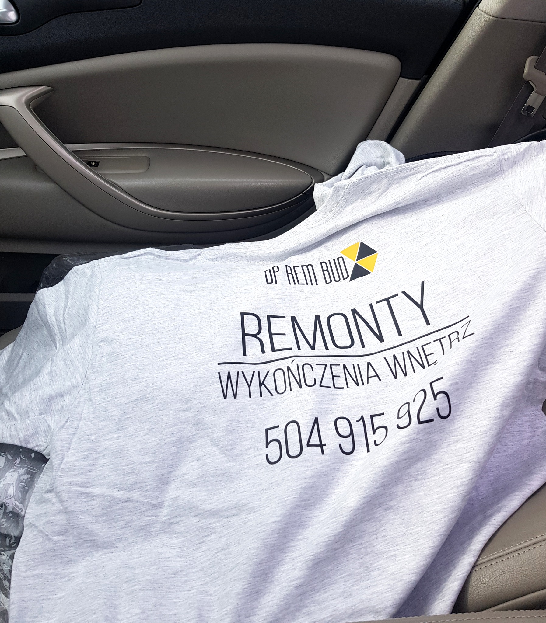 Szara koszulka z nadrukiem 'OP REM BUD Remonty Wykończenia Wnętrz 504 915 925' leżąca na siedzeniu samochodowym w Warszawie.
