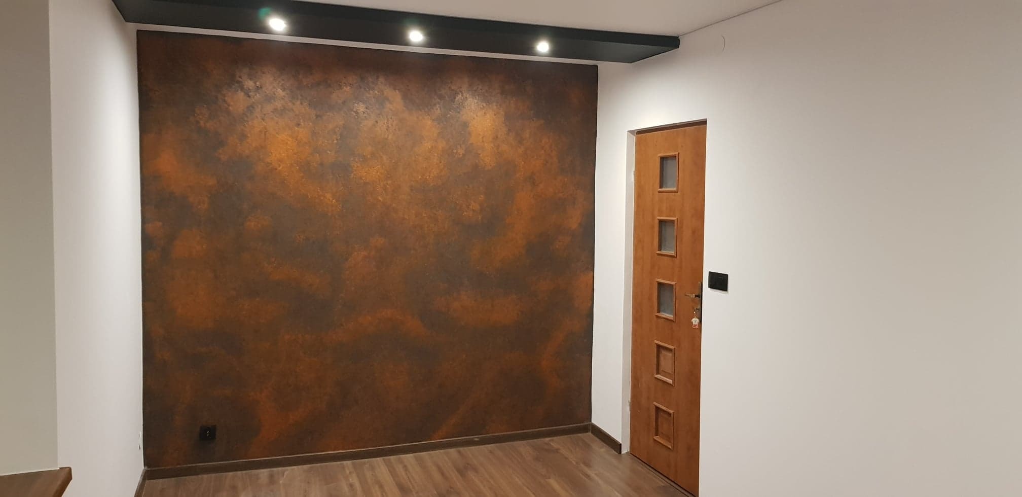 Ściana z dekoracyjną, rdzawą fakturą, oświetlona punktowymi lampami sufitowymi, sąsiadująca z białą ścianą i drewnianymi drzwiami z przeszkleniami.