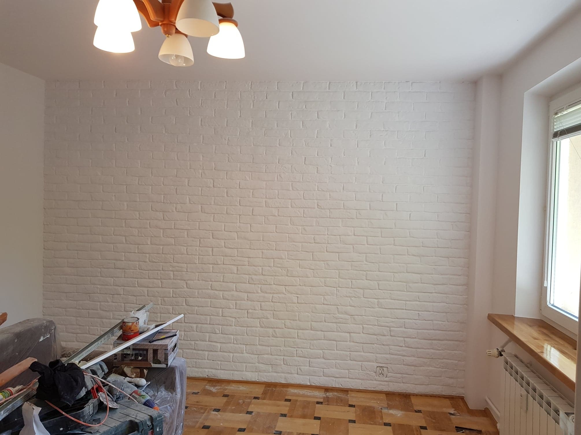 Ściana z cegieł pomalowana na biało w pokoju z parkietem, widoczne narzędzia malarskie i drabina malarska.
