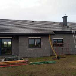 AS-DACH - Dom w trakcie ocieplania, widoczne szare płyty izolacyjne na ścianach, nowa grafitowa dachówka ceramiczna na dachu, komin z elementami drabinki, rusztowania i drabina oparta o budynek, żółty...