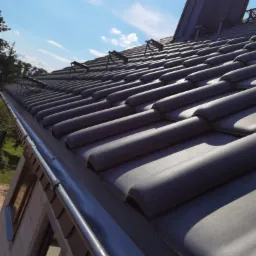 Nowy dach pokryty ciemnoszarą dachówką ceramiczną, widok z dołu na rynny i fragment ściany budynku w surowym stanie, błękitne niebo z chmurami w tle.