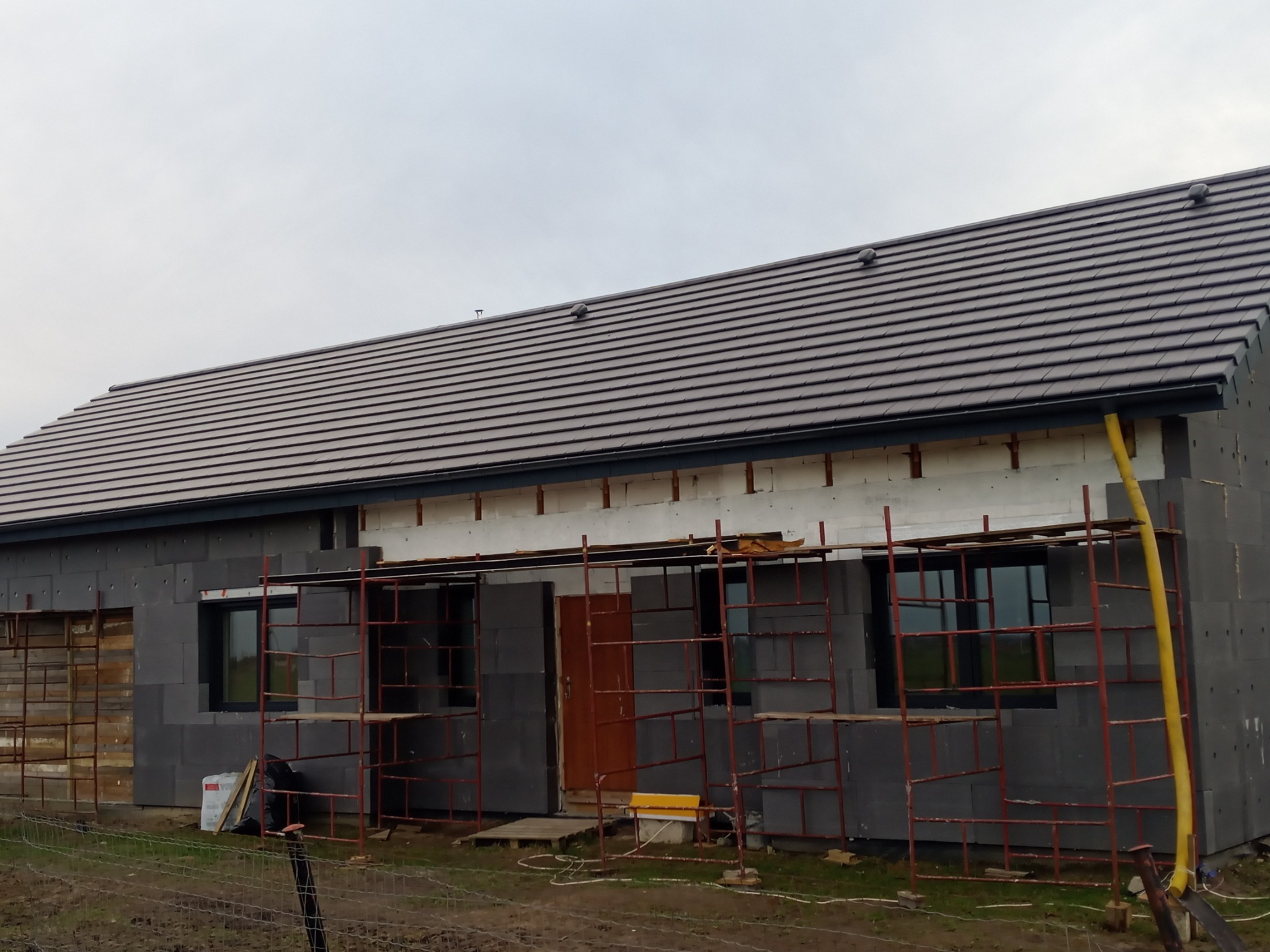 Dom jednorodzinny w trakcie budowy, widoczna elewacja z szarego styropianu, częściowo ocieplona, z zamontowanymi oknami i brązowymi dachówkami na dachu, rusztowania przy ścianach.