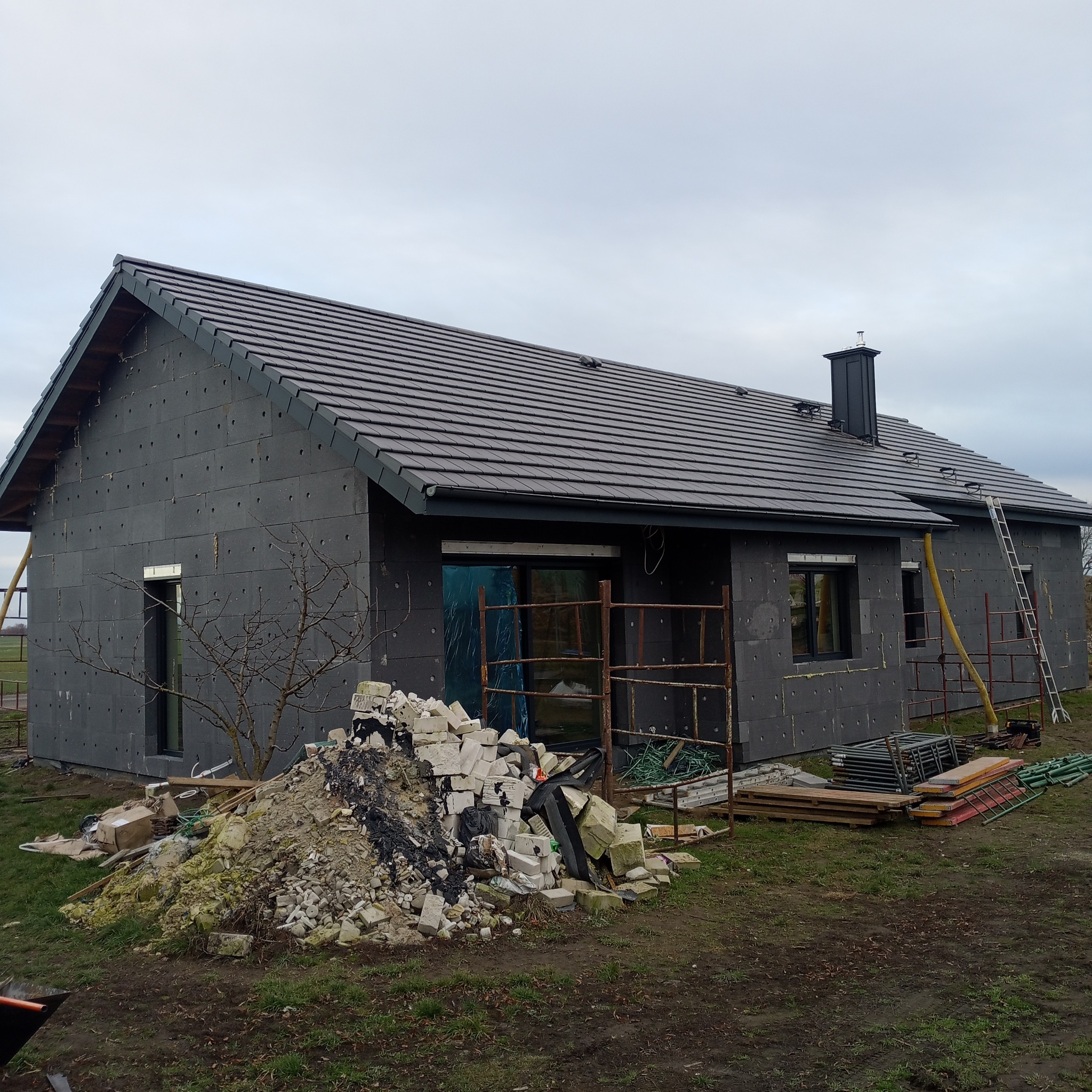 Dom w trakcie ocieplania i krycia dachu dachówką ceramiczną, widoczne rusztowania, styropian na ścianach i komin.