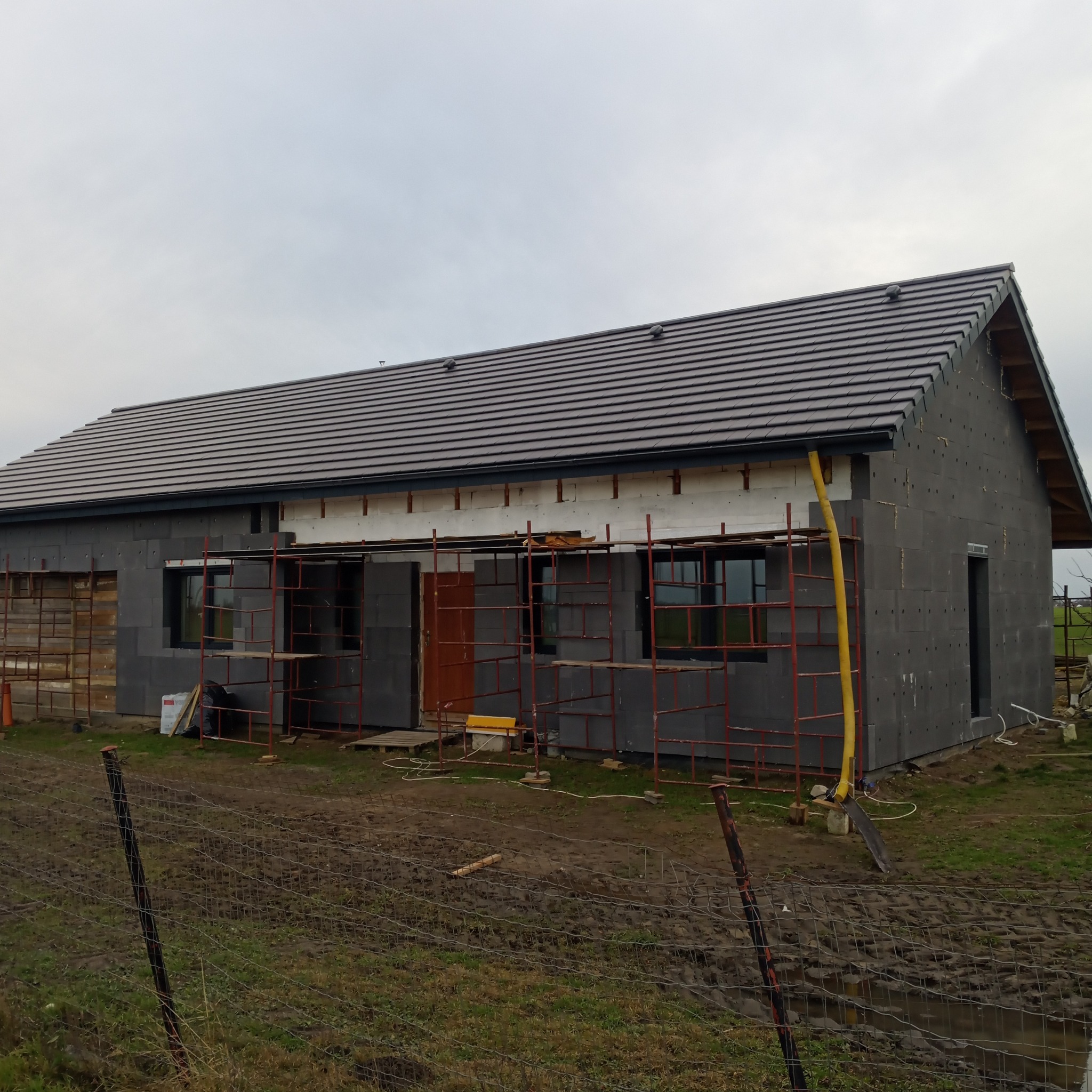 Dom w trakcie budowy, ocieplony szarym styropianem, z częściowo położonym ciemnym dachem z dachówki ceramicznej, widoczne rusztowania i teren budowy z błotem.