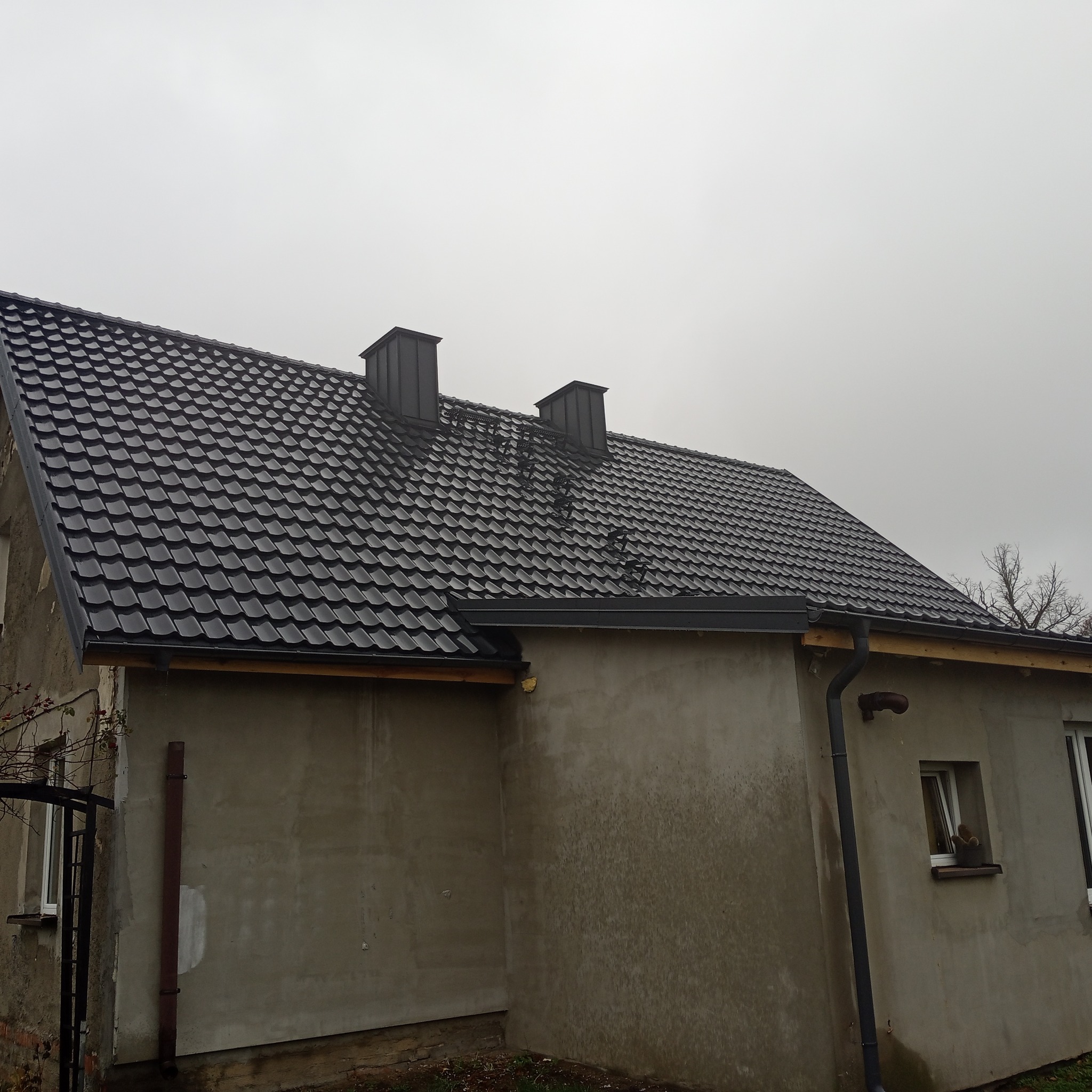 Dom jednorodzinny z nowym, ciemnym dachem z dachówki metalowej, widoczne kominy i drabinka dachowa, elewacja budynku w stanie surowym.