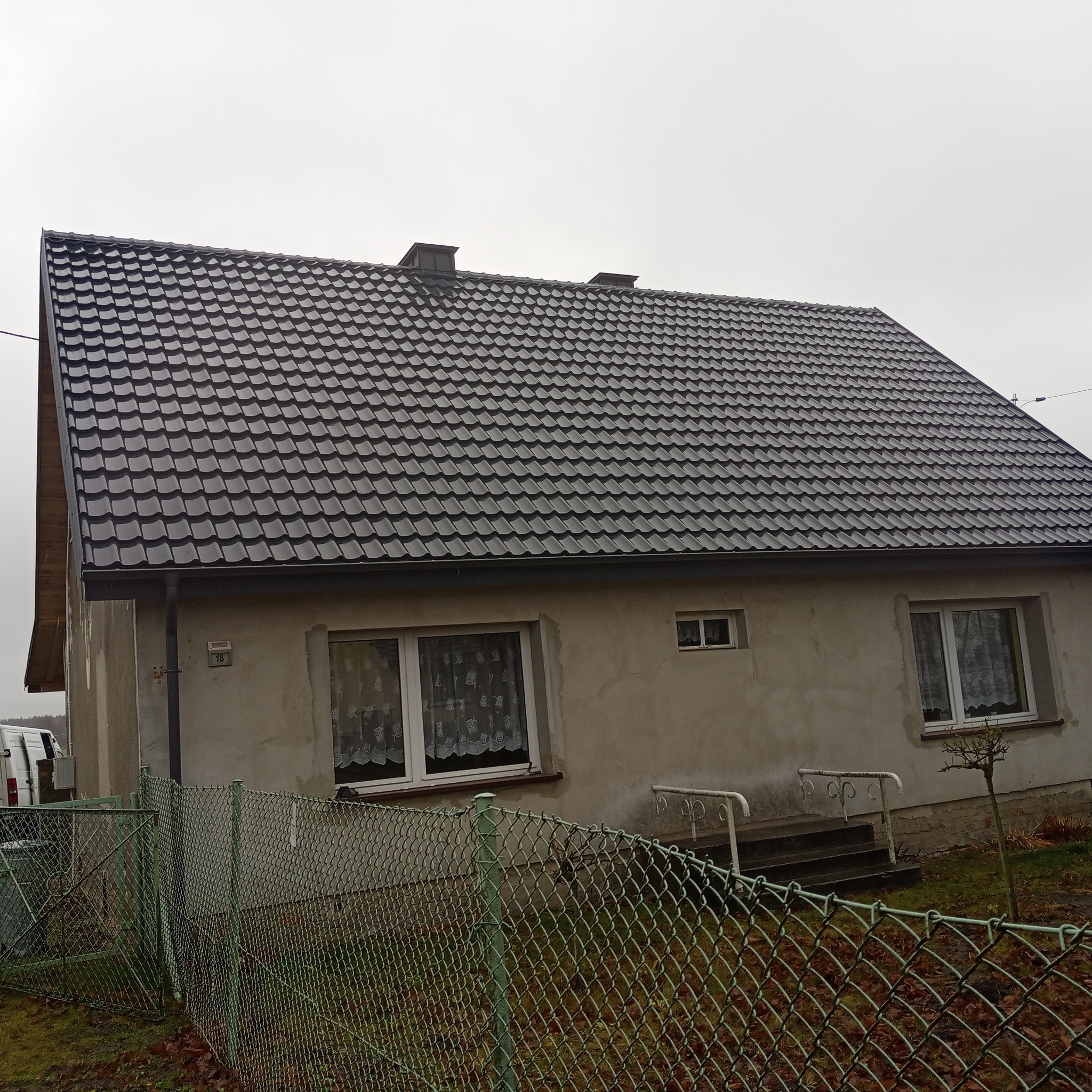 Dom w Kcyni z nowym, ciemnoszarym dachem z dachówki metalowej, widoczny zza ogrodzenia z siatki.