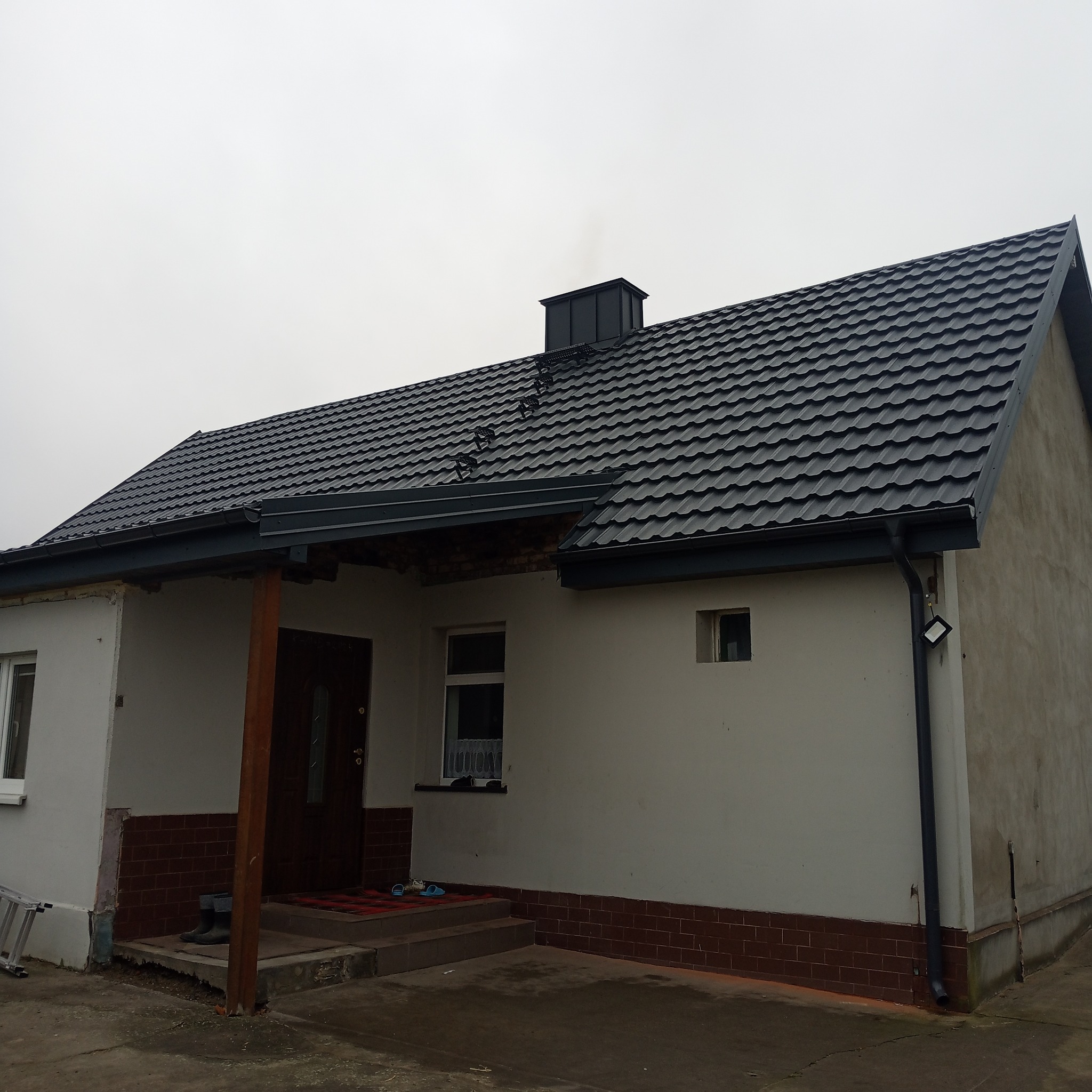 Dom jednorodzinny z nowym, ciemnoszarym dachem z blachodachówki, widoczny komin z dymem, orynnowanie i fragment elewacji w trakcie wykończenia.