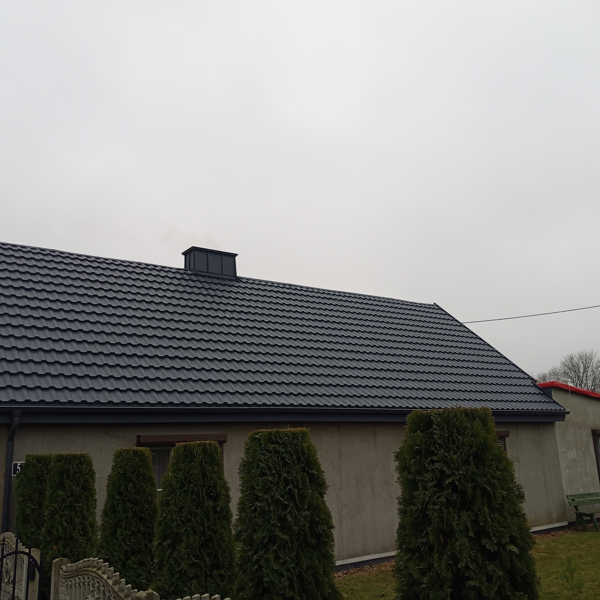 Dom jednorodzinny z nowym, ciemnoszarym dachem z dachówki ceramicznej, widoczny komin i rząd tuj przy ogrodzeniu