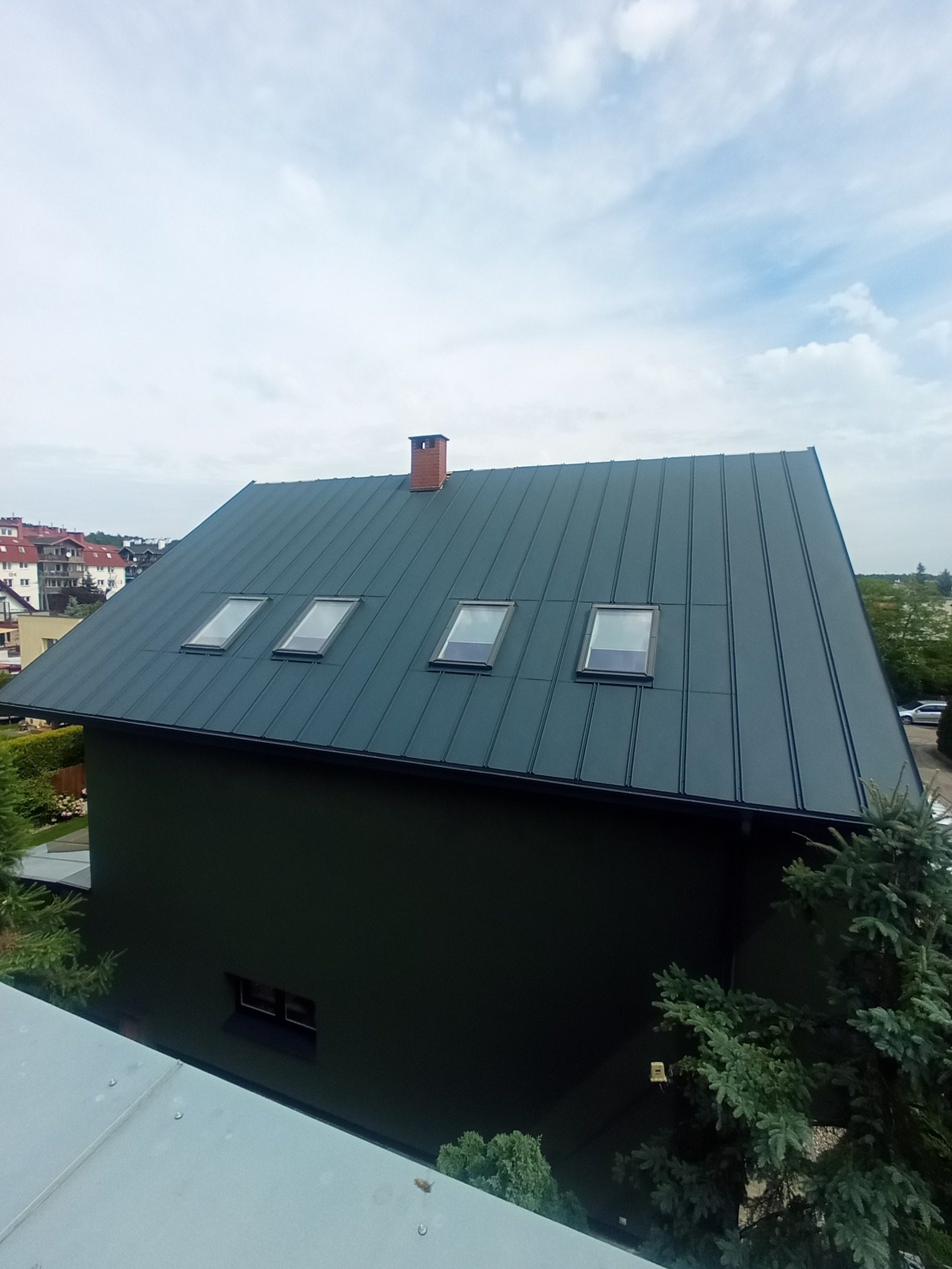 Ciemny, blaszany dach z trzema oknami połaciowymi i ceglanym kominem na tle pochmurnego nieba. Widok z góry.