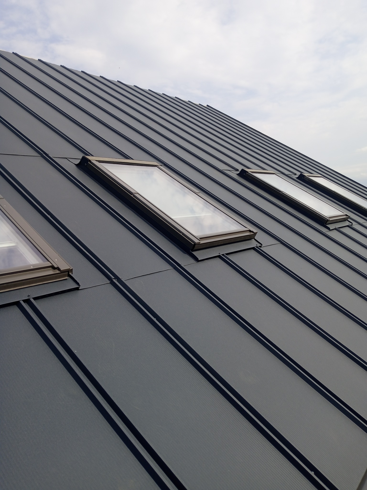 Grafitowy dach z blachy na rąbek stojący, z czterema oknami dachowymi. Widok z dołu, pochmurne niebo w tle. Minimalistyczny styl.