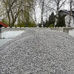 Góra szarego kruszywa drobnoziarnistego za betonowym murem, w tle brzozy i budynek mieszkalny pod pochmurnym niebem.