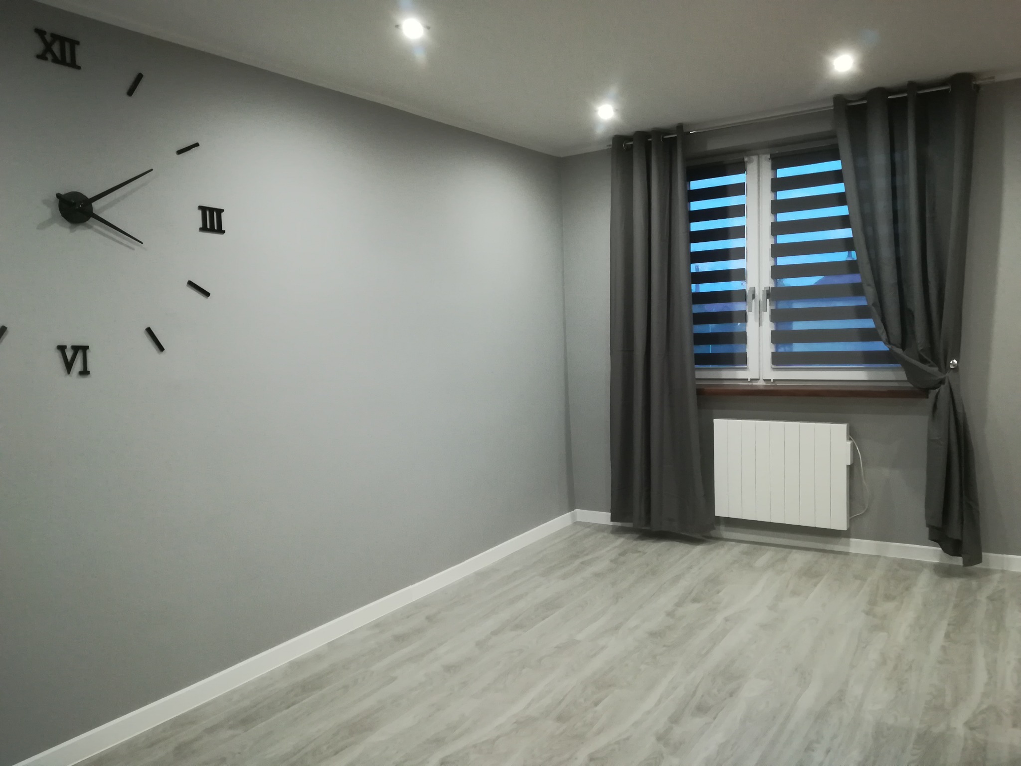 Szare wnętrze pokoju po remoncie: ściany pomalowane na szaro, panele podłogowe imitujące drewno, biały grzejnik pod oknem z szarymi zasłonami i duży zegar ścienny z rzymskimi cyframi.