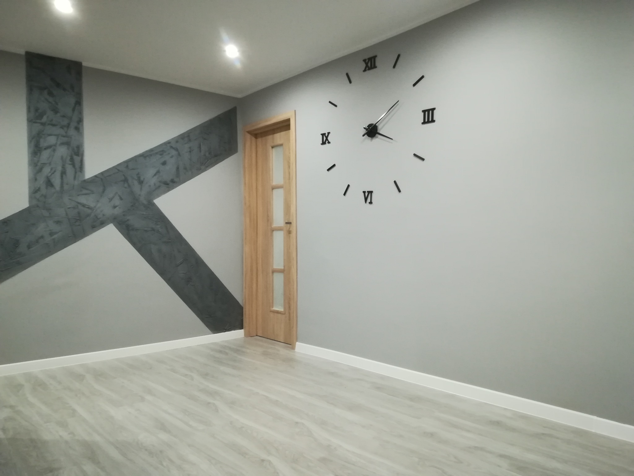 Szare wnętrze pokoju z drewnianą podłogą, ścianą z dekoracyjnym szarym wzorem w kształcie litery X, duży czarny zegar rzymski na ścianie i drewniane drzwi z szybą.
