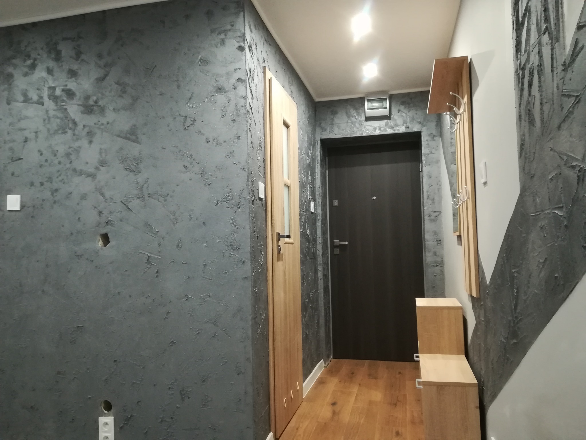 Korytarz po remoncie z szarą ścianą o dekoracyjnej fakturze, drewnianą stolarką drzwiową i minimalistycznym wieszakiem z szafkami.