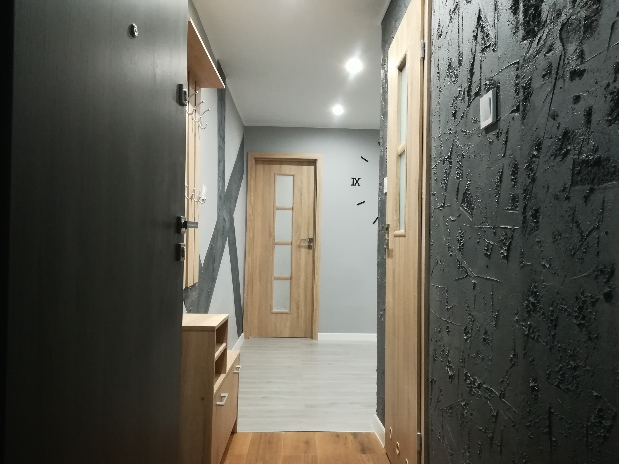 Wykończony korytarz z ciemną teksturowaną ścianą, drewnianymi drzwiami i szafką na buty w jasnym drewnie, widok na dalszą część mieszkania z szarą ścianą i dekoracyjnym zegarem.