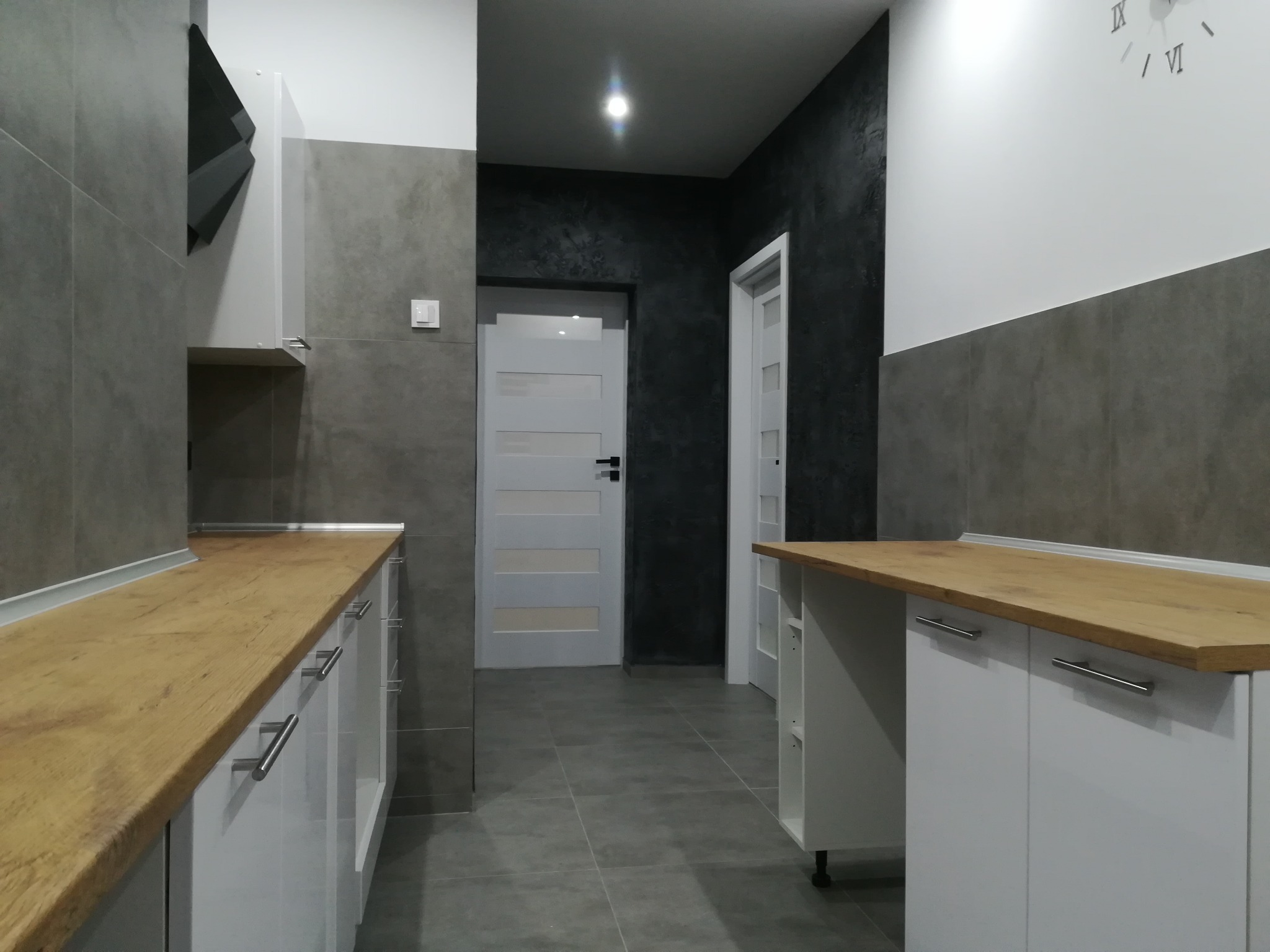 Nowoczesna kuchnia z szarymi płytkami, drewnianym blatem i białymi szafkami, widoczne drzwi wewnętrzne z poziomymi panelami i zegar na ścianie.