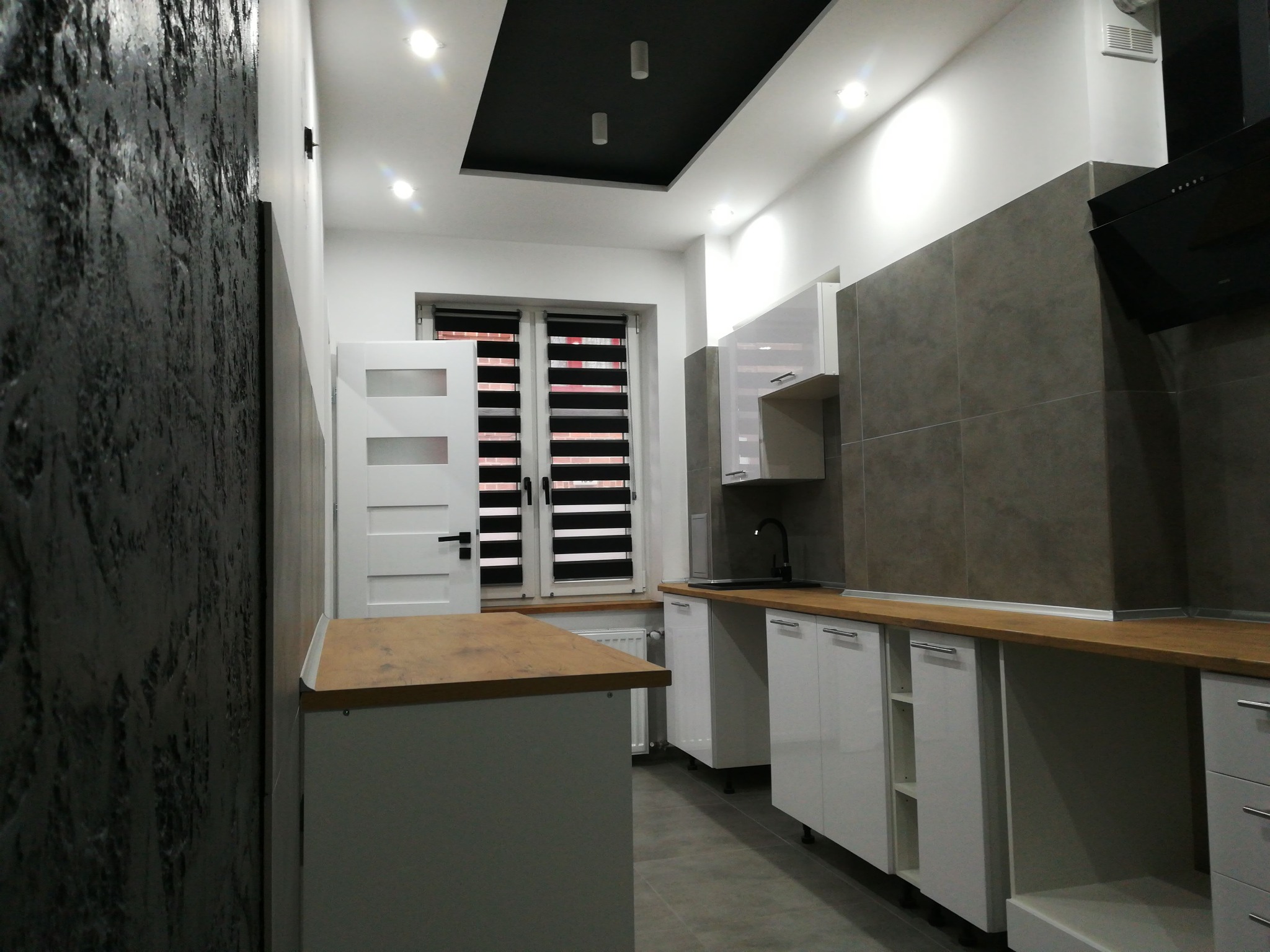 Nowoczesna kuchnia z wyspą, ściana z fakturą betonu, białe szafki z drewnianym blatem, czarny sufit podwieszany z oświetleniem punktowym.